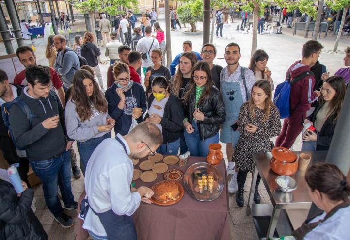 Estudiantes del Grado en Gastronomía de la UCAM, en primer plano, dando a probar las elaboraciones preparadas, mientras al fondo, se disputan los torneos de esgrima y ajedrez.